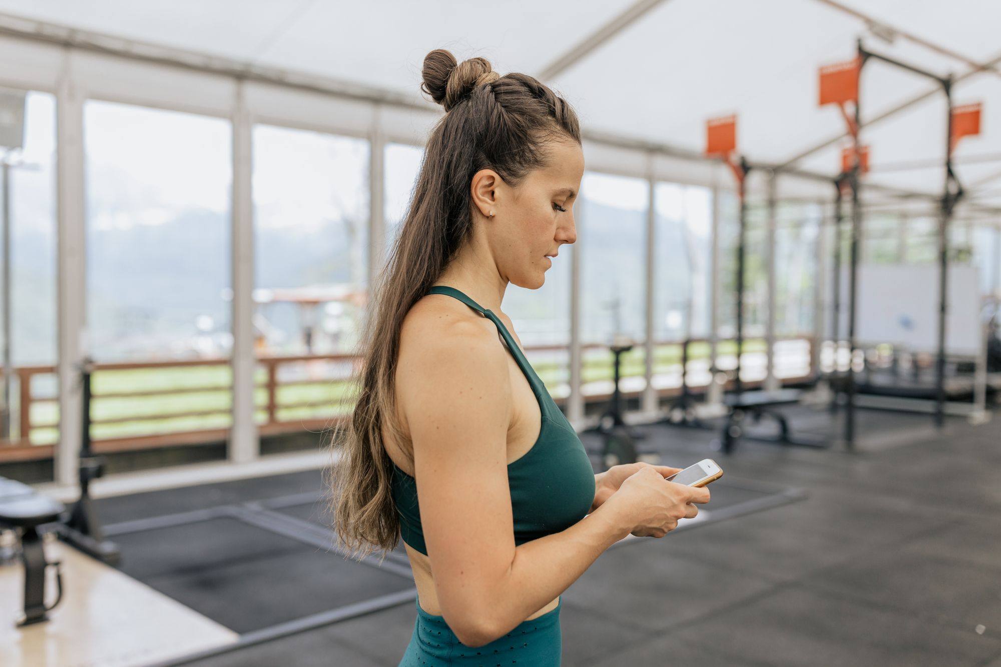 Mujer con ropa deportiva escribiendo en su móvil en un gimnasio luminoso con racks al fondo