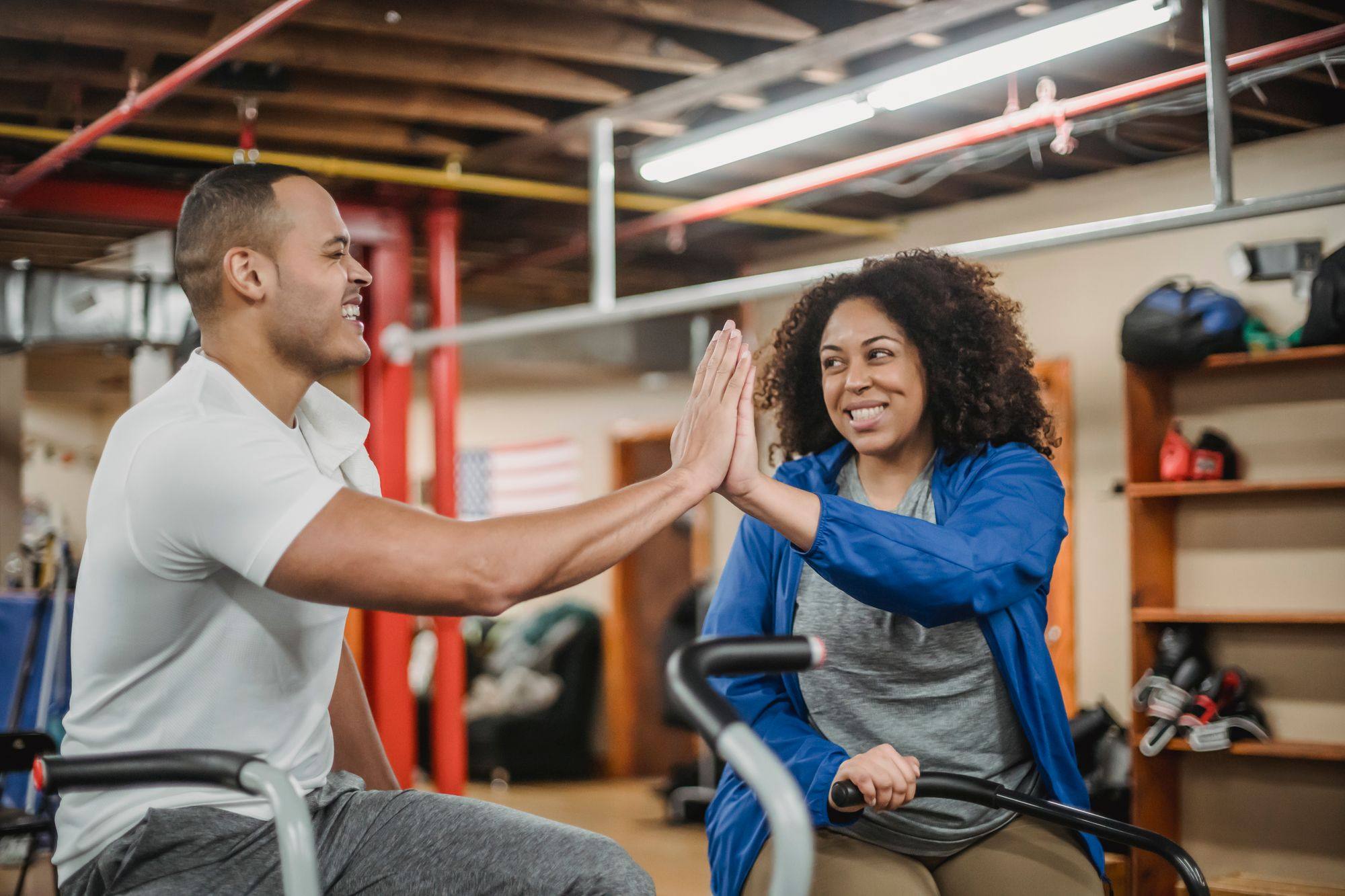 Dos personas sonríen y chocan la mano sobre bicicletas estáticas en un gimnasio de boxeo
