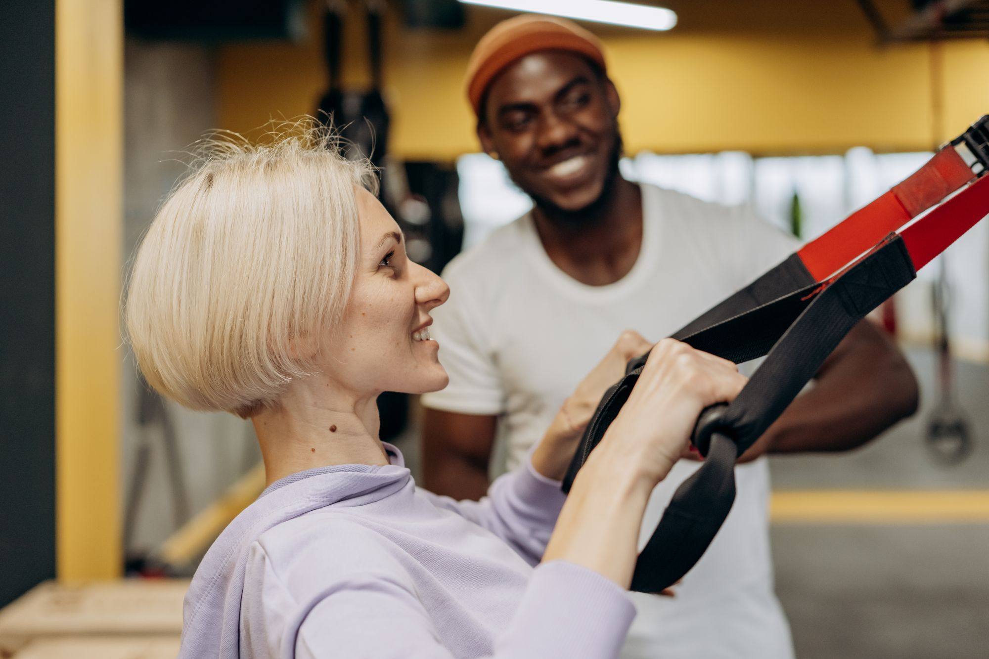 Clienta sonriente entrena con cintas de suspensión y charla con su entrenador en el gimnasio